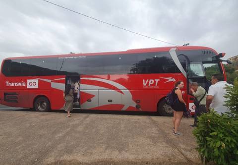 Red tour bus with passengers boarding beside rural road on cloudy day.