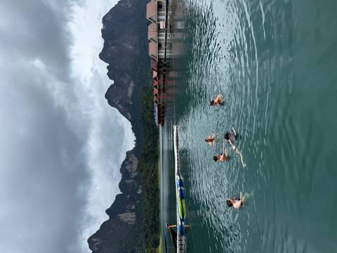 Travelers swimming in a calm emerald lake bordered by dramatic limestone mountains and floating bungalows.