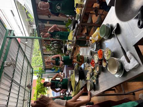 Participants wearing aprons gather around stainless tables during a Thai cooking class with fresh ingredients laid out.
