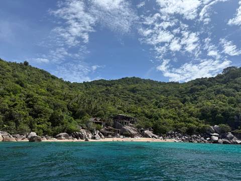 Lush green hillside meets turquoise sea and a secluded sandy beach under a bright blue sky with wispy clouds.