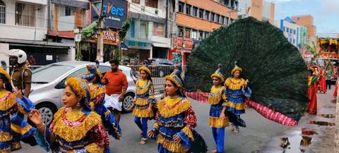 Colorful street parade with costumed children and a giant peacock feather display.