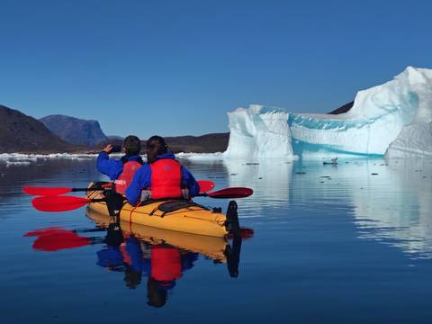 Two kayakers paddle past a towering iceberg reflected in calm Arctic waters.