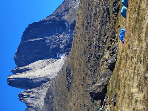 Steep granite spire dominates alpine landscape with tiny blue tents at its base.