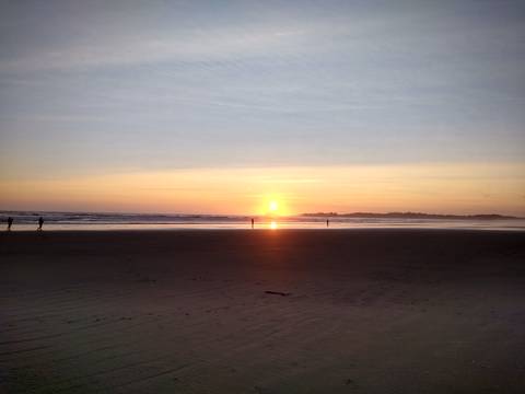 Sun sets over a wide sandy beach with a few silhouetted figures near the shoreline.