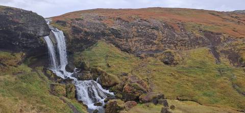 Waterfall cascades over dark rocks into a mossy ravine amid autumn-coloured hills.