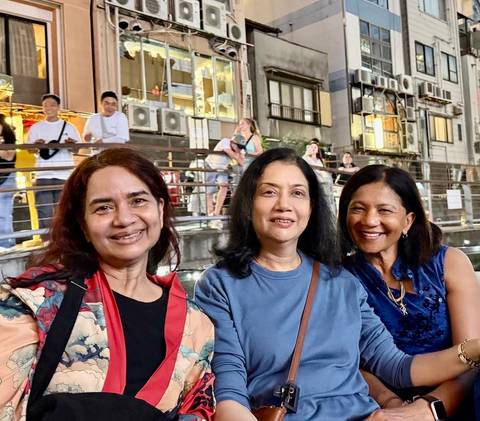 Three women smile together along a lively riverside promenade at night.