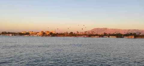 Early morning light bathes the Nile with distant balloons over a riverside town and desert mountains.