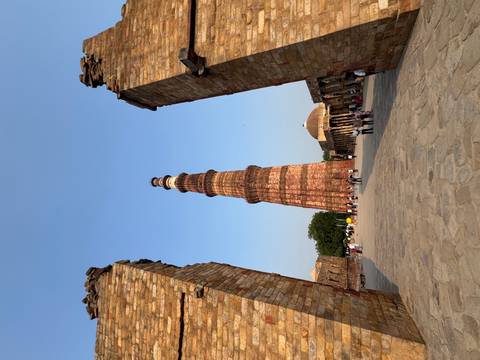 Tall red-sandstone Qutub Minar framed by ruined gateway pillars with visitors below.
