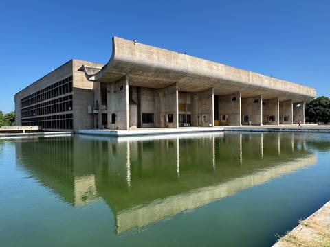 Modern concrete government building in Chandigarh reflected perfectly in a still green pool beneath a bright blue sky.
