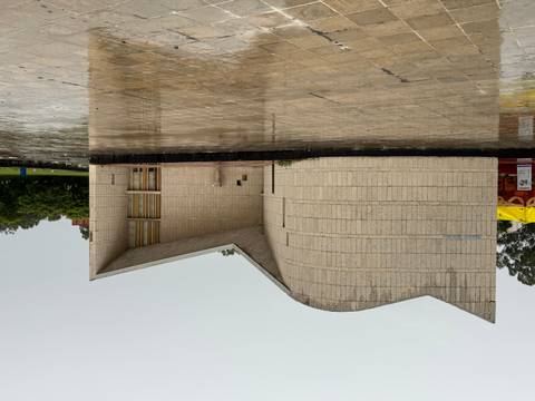 Small curved concrete museum building on a rain-soaked plaza under grey skies.