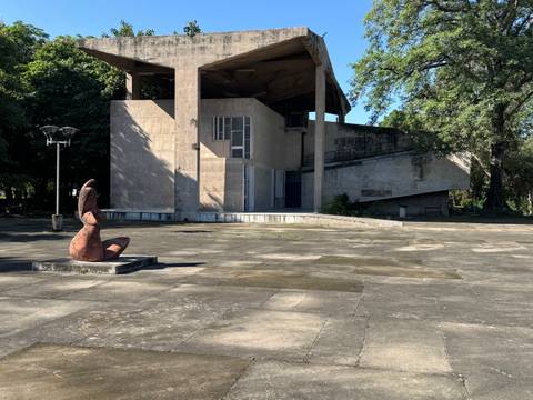 Open concrete pavilion with a seated abstract sculpture in a spacious plaza shaded by trees.