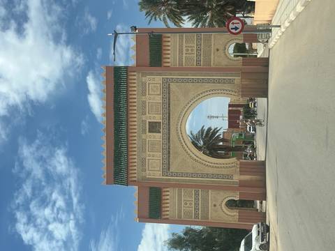 Tall ornamental archway with geometric tilework marks the entrance to a desert town under a partly cloudy sky.