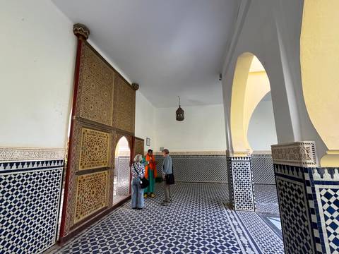Visitors listen to a guide in a tiled chamber lined with intricate Moroccan mosaics and carved wooden doors.