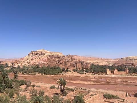 UNESCO-listed mudbrick ksar of Ait Benhaddou rises above a dry riverbed and sparse palms beneath a clear sky.