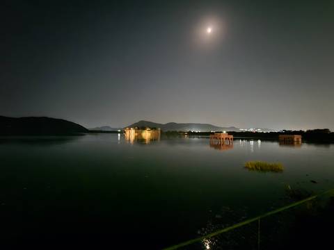 Nighttime long-exposure of Jal Mahal palace reflecting on a calm lake under a faint moon.
