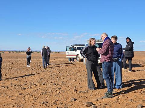 Travel group chats beside 4×4 vehicles on a wide, stony desert expanse under clear skies.