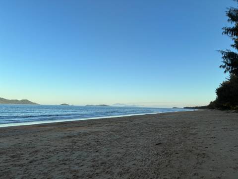 Empty sandy beach with calm blue sea and distant forested headlands under a clear sky.