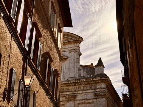 Warm sunlight illuminates brick facades and a classical church viewed from a narrow Italian alley.