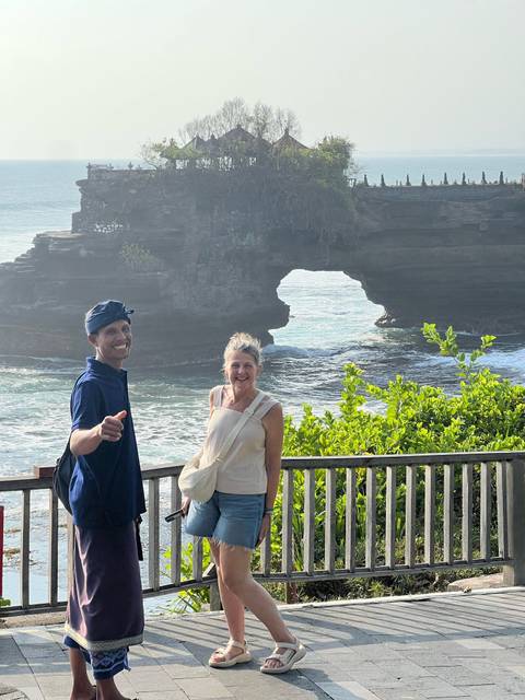 Two travellers smiling in front of an ocean rock arch and temple cliffs with lush greenery.