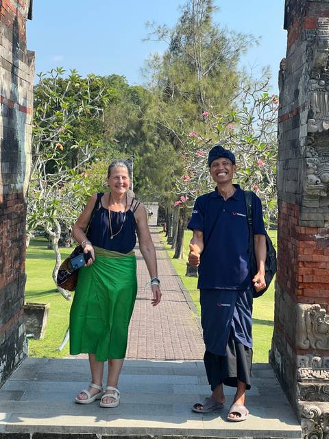 Two smiling travellers framed by an ornate Balinese split gate and flowering trees.