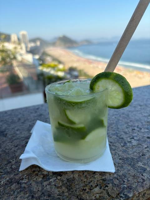 Close-up of a chilled caipirinha cocktail garnished with lime against a blurred beach backdrop.