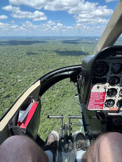 View from helicopter cockpit over dense green rainforest with flight instruments visible.