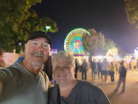 Nighttime selfie of a smiling couple at a fairground with a colorful illuminated ferris wheel behind them.