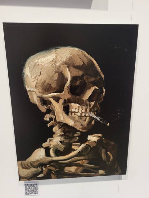 Photograph of Van Gogh's painting 'Skull with Burning Cigarette' displayed on a dark wall.