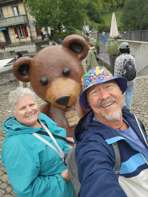Cheerful couple taking a selfie with a large teddy bear statue in a cobblestone square.