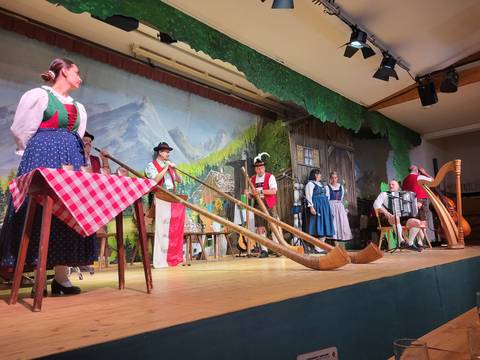 Traditional alpine show with performers playing long alphorns on stage before a painted mountain backdrop.