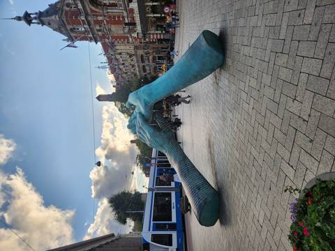 Large turquoise sculpture of two hands touching in a busy Amsterdam square with trams passing.
