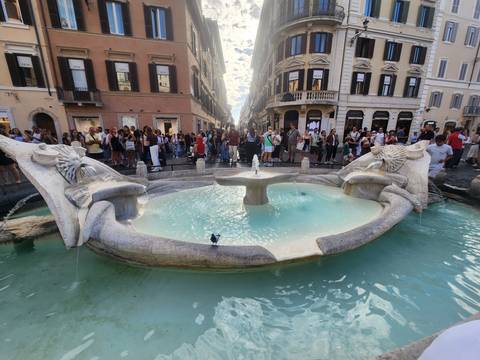 Crowds gather around the turquoise water of the Barcaccia Fountain at the foot of elegant Roman streets.