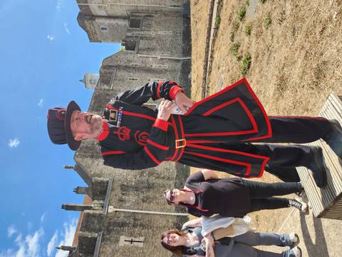 Uniformed Yeoman Warder in black and red stands before the stone walls of the Tower of London.
