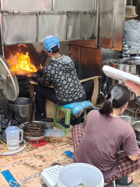 Street-food vendors grill skewers over roaring flames amid stainless steel pots and fans.