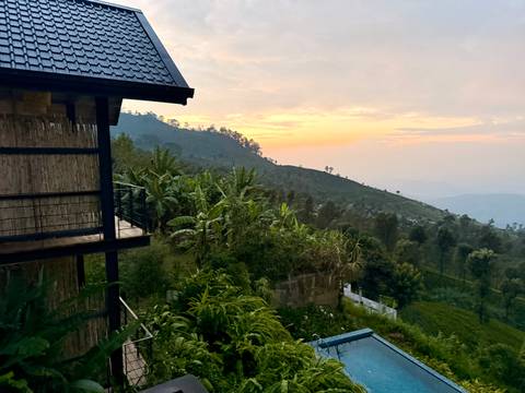 Serene hill-country landscape at sunset viewed from a wooden balcony, with banana trees below.