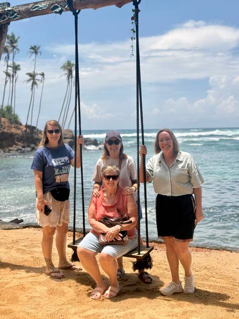 Women posing on a swing beside the ocean with waves crashing behind.
