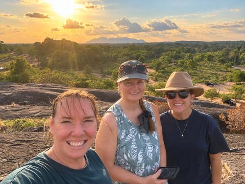 Three women smiling on a rocky viewpoint at golden sunset with forested plains and distant hills behind them.