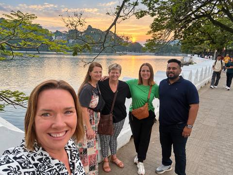 Five travelers standing by a tranquil lakeside promenade at sunset with town lights beginning to glow.