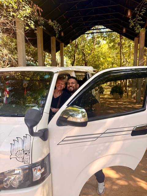 Traveler and driver smiling from the front seats of a white touring van parked under trees.
