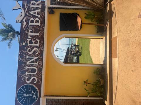 Yellow-walled entrance archway marked 'SUNSET BAR' framing a lawn and beachside seating with palm trees and ocean beyond.