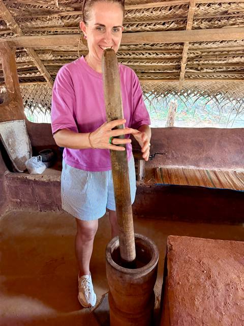 Person in casual clothes using a long wooden pestle inside a rustic mud-brick hut with thatched roof.