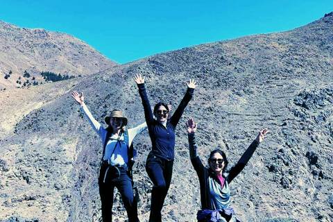 Three hikers with arms raised in triumph on rocky High Atlas mountain ridge under clear blue sky.