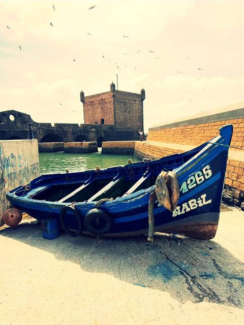 Blue wooden fishing boat moored beside historic stone ramparts and arches of Essaouira harbor.