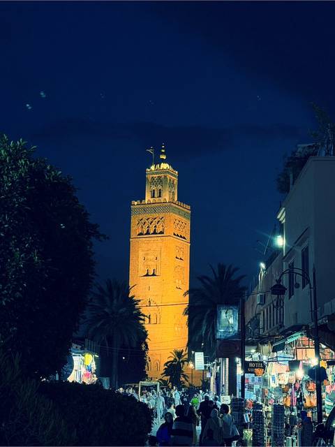 Night view of the illuminated Koutoubia Mosque minaret rising above palm trees and dark blue sky.