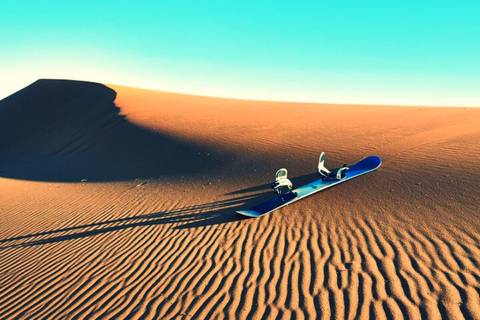 Isolated snowboard resting on rippled sand at the base of a tall desert dune under clear sky.