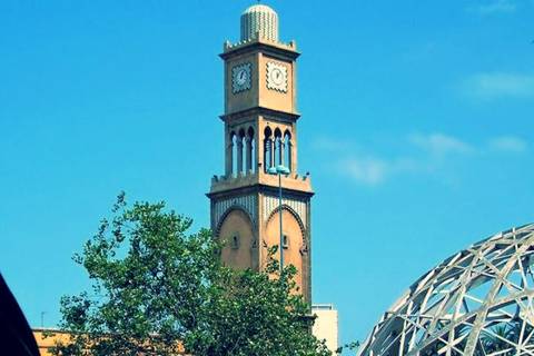 Historic yellow clock tower rising beside modern glass dome against a clear blue sky.