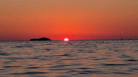 Fiery red sun setting into calm Adriatic Sea with small island silhouette on horizon.