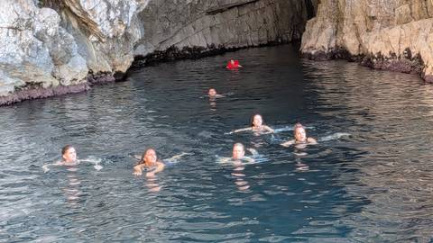 Travelers swimming in deep blue water inside a rocky sea cave entrance.