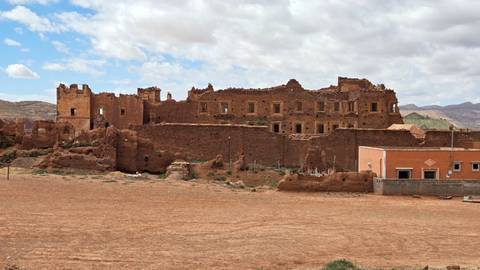 Ruined earthen kasbah walls and empty windows stand in a barren landscape under a partly cloudy sky.
