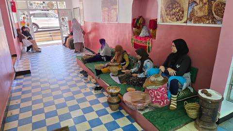 Women seated on cushions hand-grinding argan nuts inside a cooperative with tiled blue-white floor.